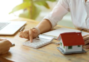 Person's finger pushing calculator button at desk with small model home nearby. Concept for manufactured home cost.