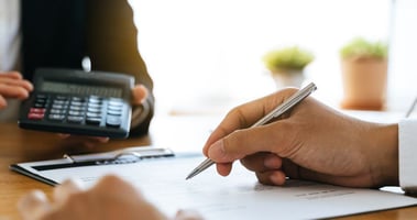 Hand signing insurance papers with calculator nearby. Signifies factors that affect home insurance prices